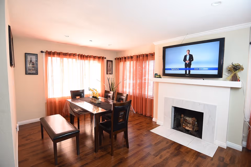 A dining area with a wooden table covered by a clear plastic tablecloth, surrounded by four chairs and a bench. The room has wooden flooring, orange curtains on the windows, a fireplace with a mounted flat-screen TV above it, and framed artwork on the walls.