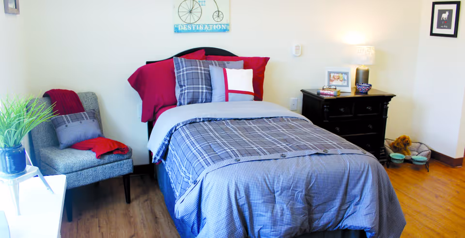 Cozy single bed in a furnished bedroom with plaid bedding, a nightstand and lamp, an armchair, and decorative accents.