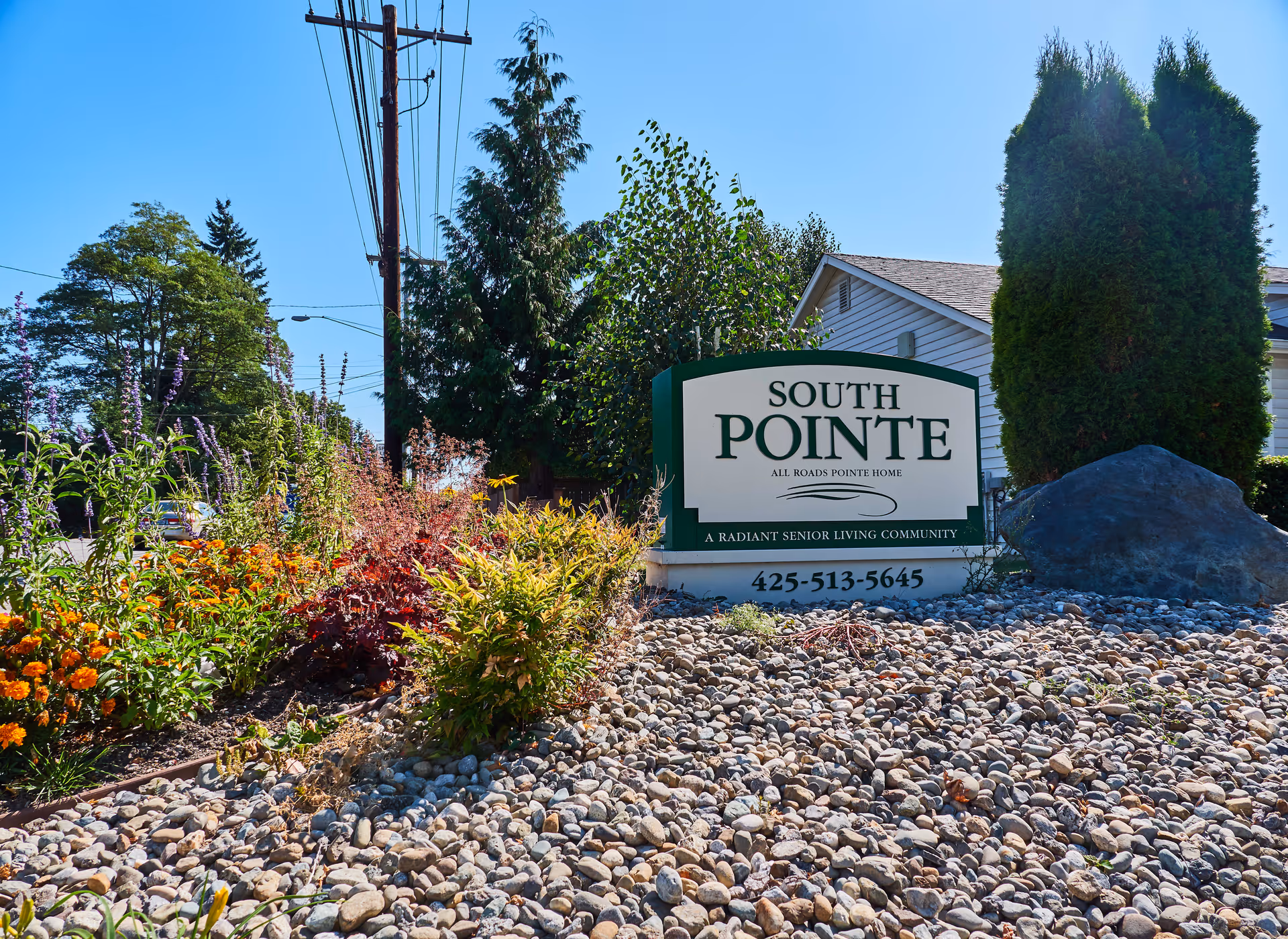 Outdoor view of the South Pointe Assisted Living facility sign surrounded by landscaping with rocks, bushes, and flowers under a clear blue sky.