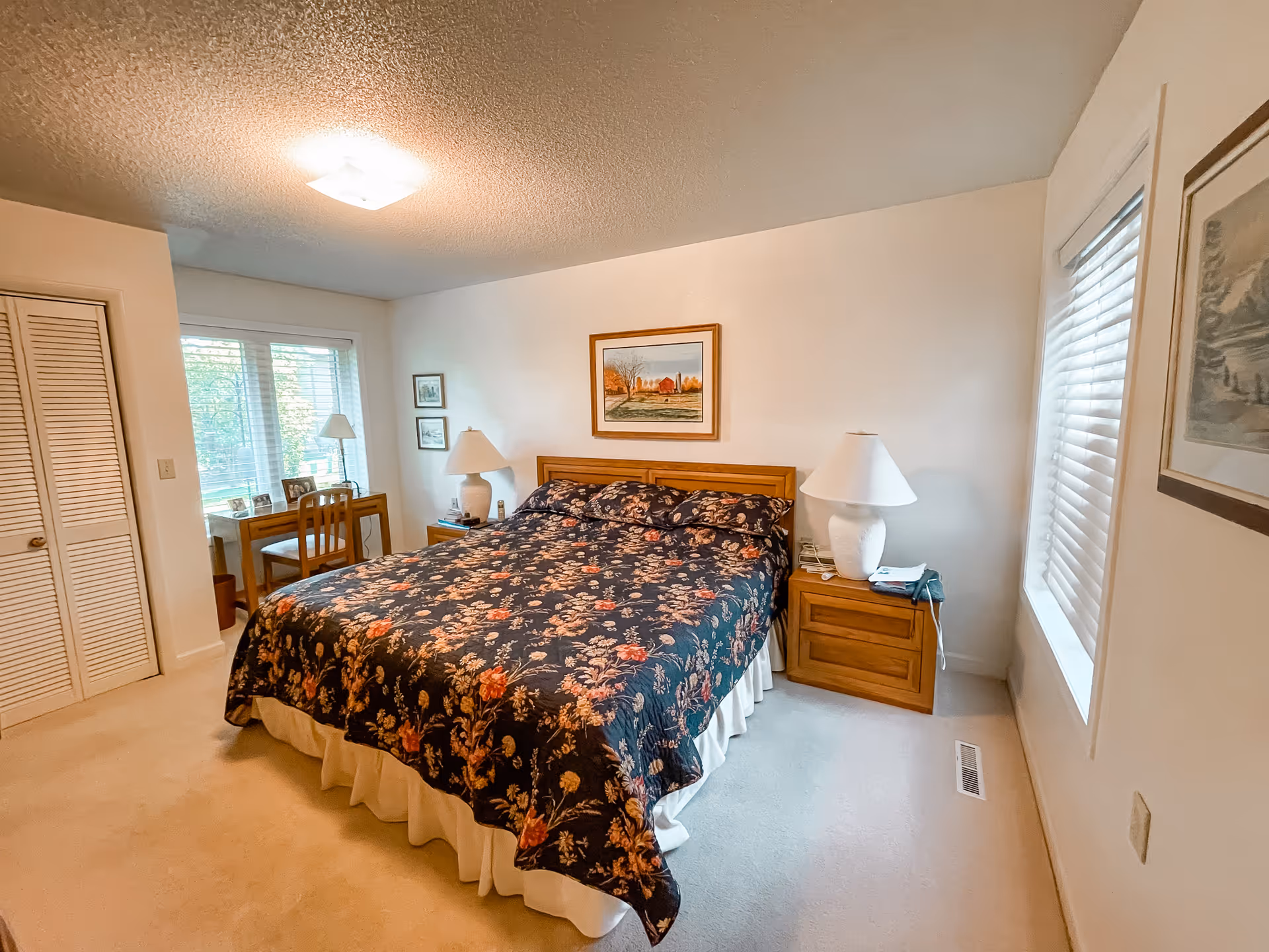 A cozy bedroom with a large bed covered in a dark floral bedspread. There are two wooden nightstands on either side of the bed, each with a white lamp. A framed painting hangs above the bed. To the left, there is a window with blinds and a small wooden desk with a chair and a lamp. The room has light-colored walls and carpeted floor.