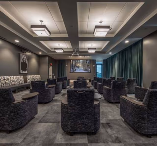 A modern media or presentation room with multiple rows of dark upholstered armchairs arranged facing a large screen at the front. The room features a patterned bench along one wall, dark curtains on the windows, and ceiling lights with rectangular fixtures.