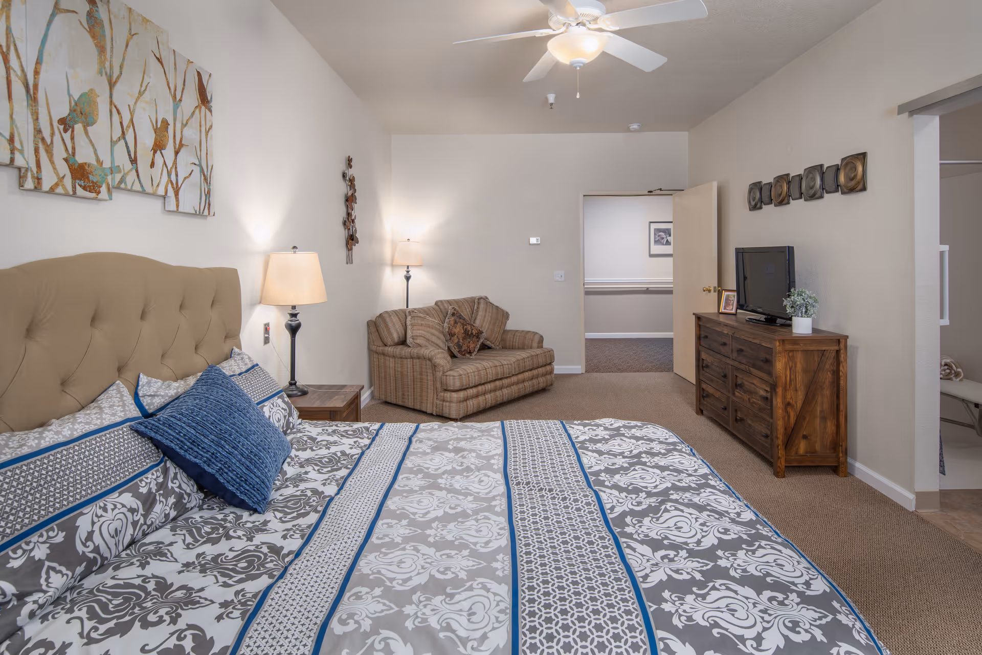 A cozy bedroom in an assisted living facility featuring a large bed with patterned bedding and blue accent pillows. There is a beige tufted headboard, a wooden nightstand with a lamp, a striped armchair with decorative pillows, a wooden dresser with a flat-screen TV and a small plant, and wall art. The room has beige walls and carpet, a ceiling fan with a light, and an open door leading to another room.
