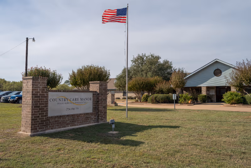 Exterior view of Country Care Manor, a skilled nursing and rehabilitation facility. The image shows a brick sign with the facility's name, an American flag on a flagpole, a parking area with cars, and the main building surrounded by trees and shrubs under a partly cloudy sky.