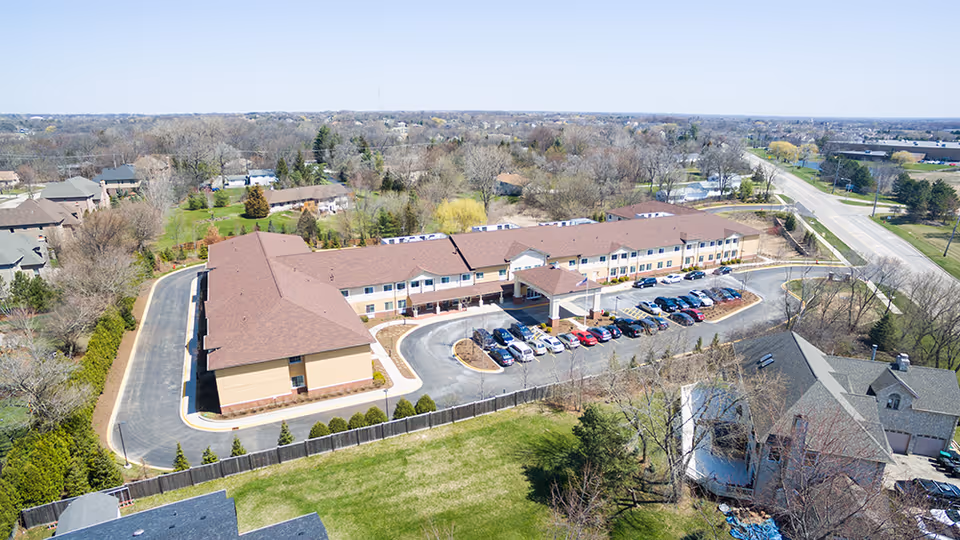 Aerial view of Cedar Lake Assisted Living and Memory Care facility showing a large two-story building with a brown roof, surrounded by parking lots with several cars, trees, and nearby residential houses under a clear sky.