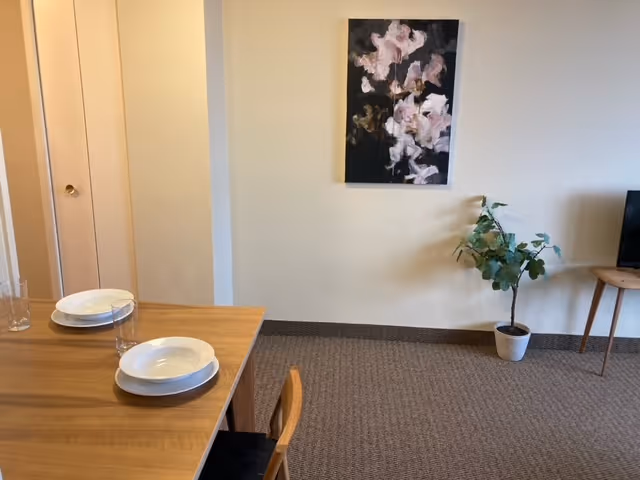 A simple interior room with a wooden dining table set with white plates and clear glasses. A wooden chair is partially visible. On the right side, there is a small potted plant on the floor and a wooden side table with a television on it. A floral abstract painting hangs on the beige wall above the plant.
