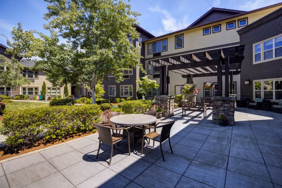 Outdoor patio area at The Lodge at Mallard's Landing featuring a round table with four chairs, a pergola with hanging flower baskets, surrounded by greenery and a multi-story building under a clear blue sky.