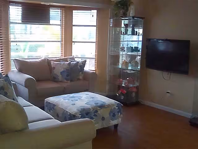 A cozy living room with two beige sofas, a floral-patterned ottoman, a glass display cabinet filled with decorative items, a wall-mounted flat screen TV, and large windows with wooden blinds letting in natural light.
