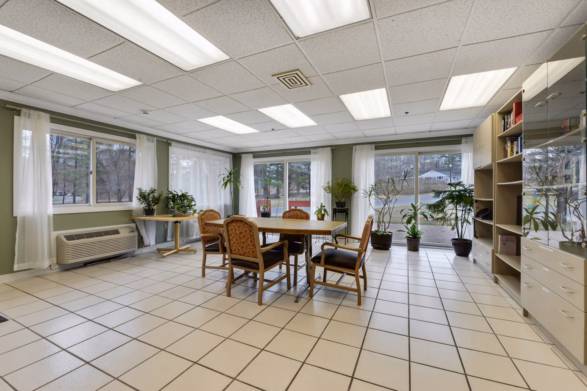 A bright room with large windows covered by sheer white curtains, featuring a wooden table surrounded by six upholstered chairs. The room has tiled flooring, several potted plants near the windows, and built-in shelving units along one wall. Fluorescent lights illuminate the ceiling.