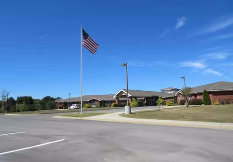 Front view of a senior living facility with flagpole, parking lot, and landscaped entrance.