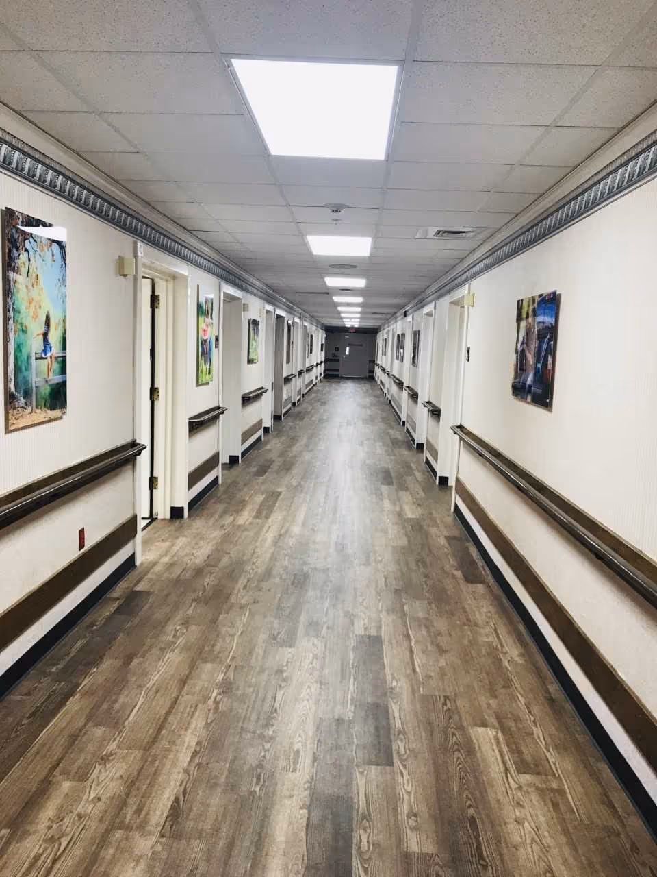 Long, well-lit hallway in a senior living facility with wood-patterned flooring, white walls, handrails on both sides, and several doors leading to rooms. Colorful framed pictures hang on the walls.