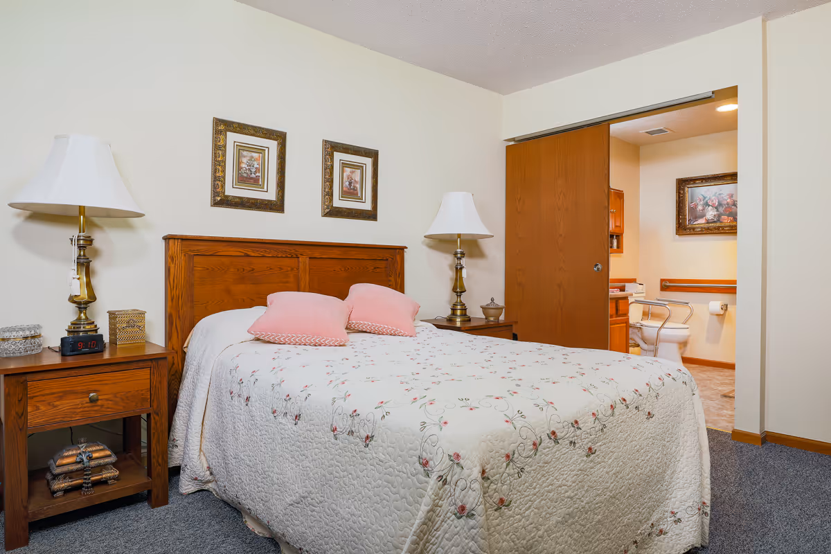 A bedroom with a wooden headboard bed covered with a white floral quilt and three pink pillows. There are two wooden nightstands on either side of the bed, each with a lamp. The nightstand on the left also has an alarm clock showing 9:10. Two framed pictures hang on the wall above the bed. An open sliding door reveals a bathroom with a toilet, grab bars, and a framed floral picture on the wall.