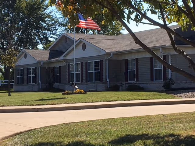 Single-story building with beige siding and dark shutters, surrounded by a well-maintained lawn and trees. An American flag is flying on a flagpole in front of the building under a clear sky.