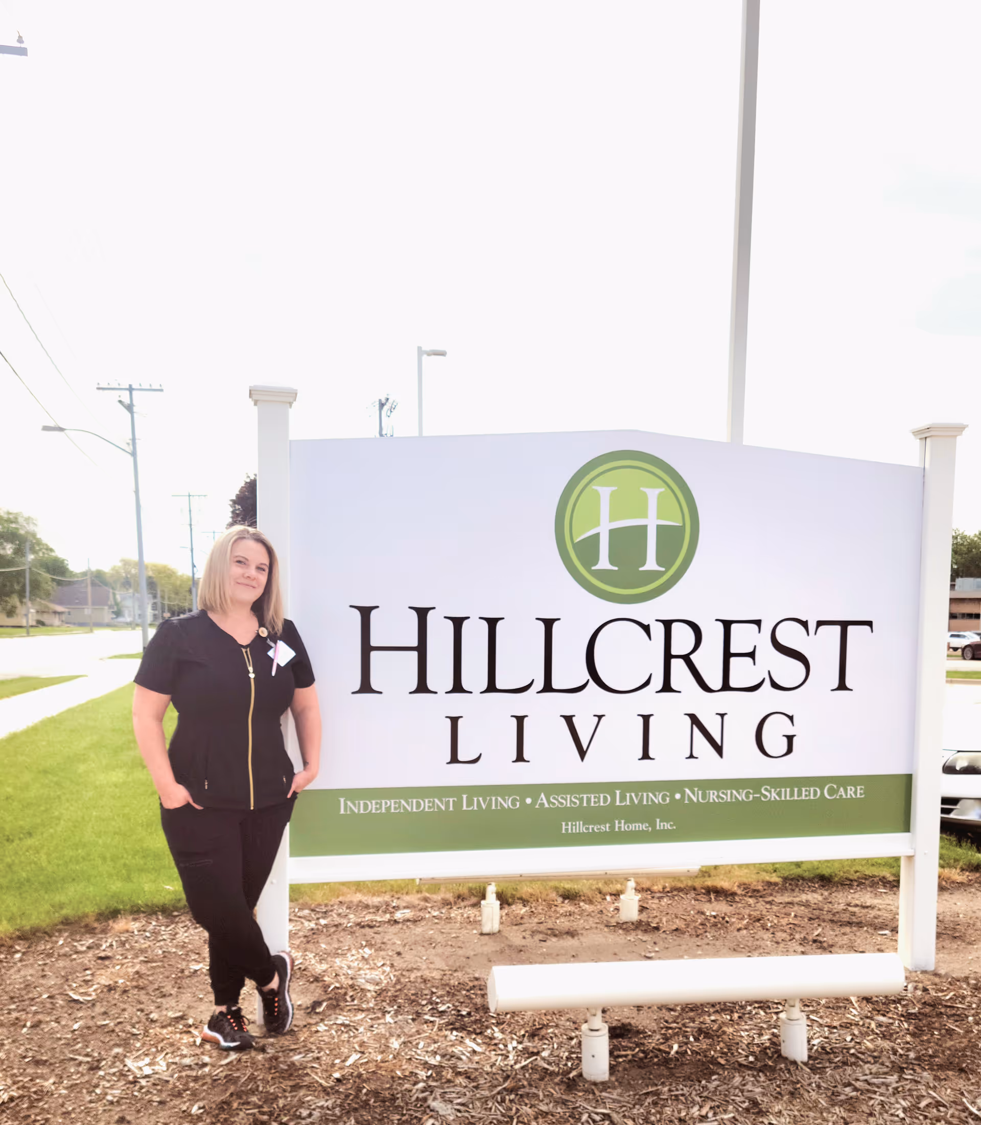 A woman stands beside a sign for Hillcrest Living Campus, which lists services like independent living and assisted living.