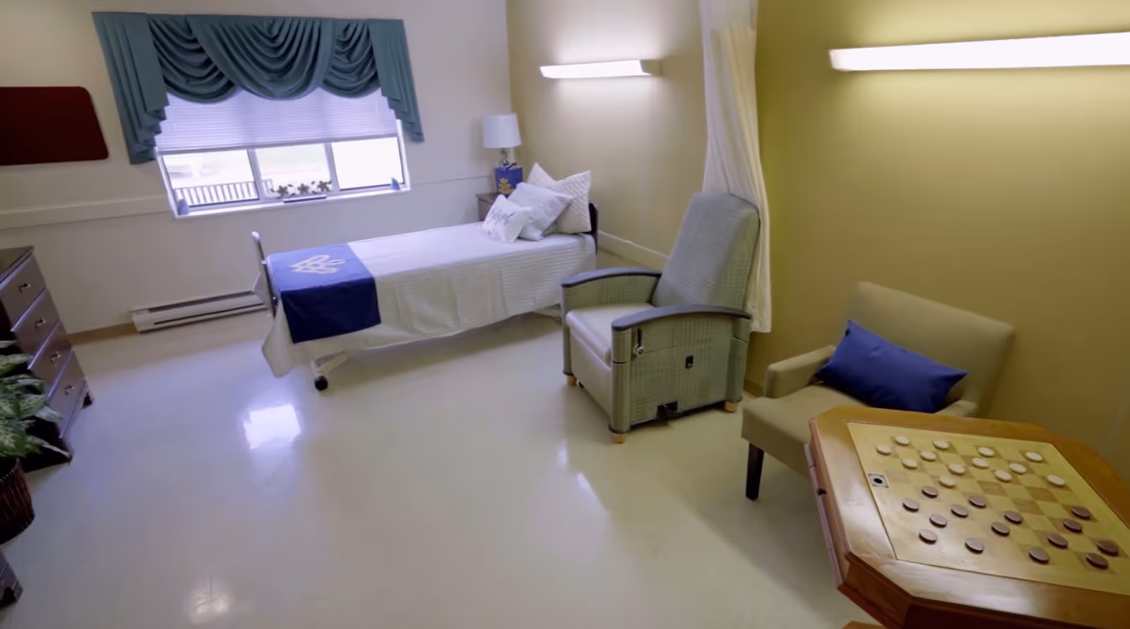 A bright and clean room with a single bed covered in white linens and a blue throw, a window with blue drapes, a bedside table with a lamp, a comfortable recliner chair, an armchair with a blue pillow, and a wooden table with a checkers board on it.