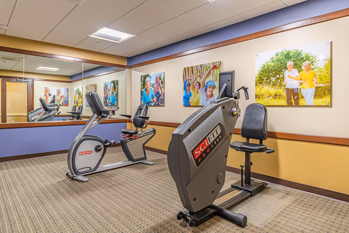 A small fitness room with two exercise bikes, one upright and one recumbent, placed on a carpeted floor. The walls are painted beige and blue with a large mirror on one side reflecting the equipment. Several framed photos of elderly people engaging in outdoor activities are hung on the walls.