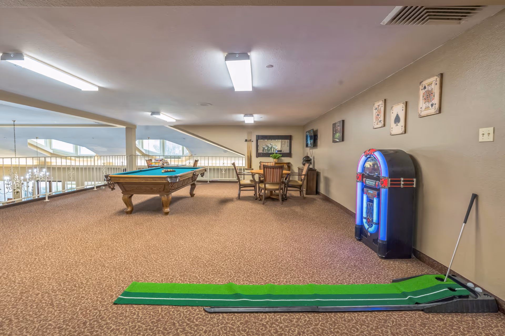 A spacious recreational room with a pool table, a small round table with four chairs, a vintage-style jukebox, and a putting green with a golf club. The room has carpeted floors, beige walls decorated with playing card artwork, and bright ceiling lights. The area overlooks a lower floor with large windows and chandeliers.
