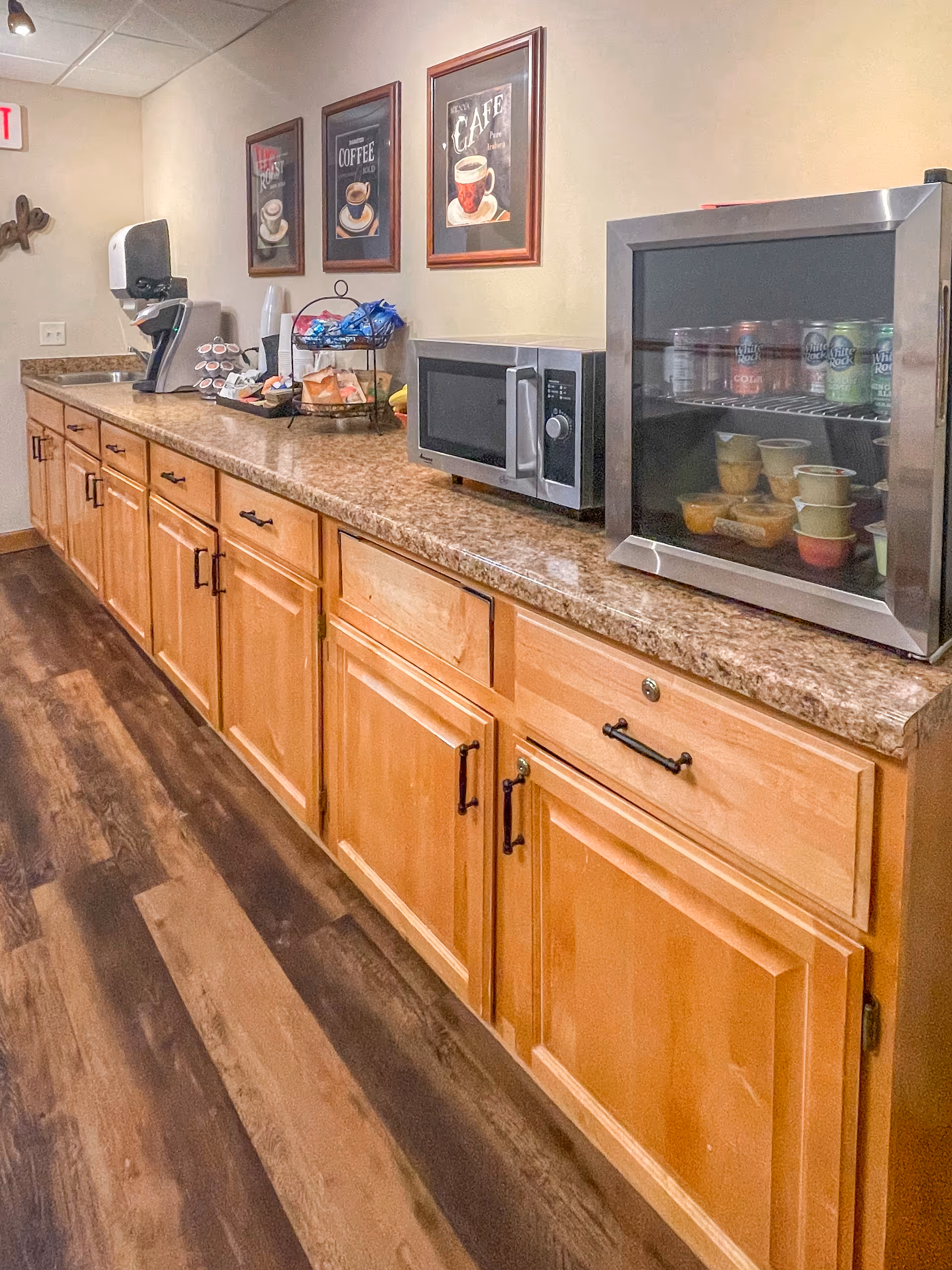 A long countertop with wooden cabinets holding a microwave, beverage cooler, coffee station and framed coffee prints on the wall.