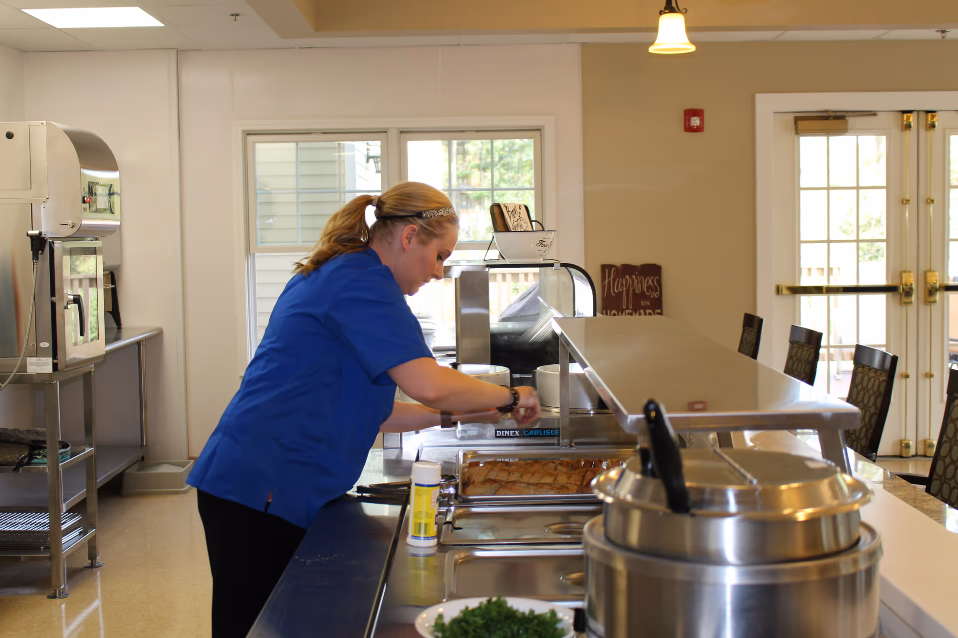A woman in a blue shirt is preparing food behind a serving counter in a kitchen or cafeteria area. There are trays of food, a large soup pot, and a container of seasoning on the counter. The room has beige walls, windows, and double glass doors in the background.