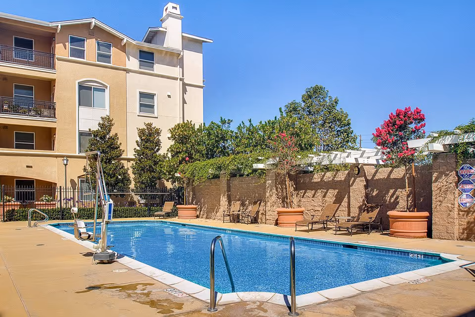 Outdoor swimming pool area at Heritage Estates with clear blue water, poolside chairs, large potted plants with flowering trees, and a multi-story beige building in the background under a clear blue sky.
