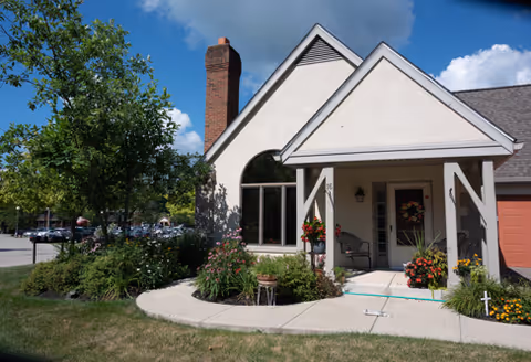 Exterior view of a single-story building with a peaked roof and chimney, featuring a covered entrance with white pillars and a door decorated with a wreath. There are flower pots with colorful flowers on either side of the entrance and a curved sidewalk leading up to it. A tree and green shrubs are visible on the left side, with a parking lot and cars in the background under a blue sky with some clouds.