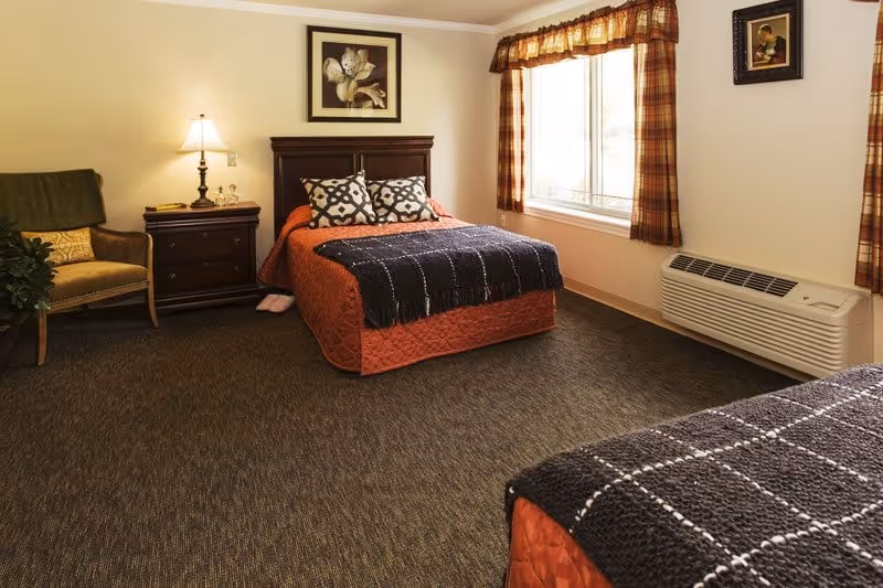 A cozy bedroom in Juniper Glen Memory Care Assisted Living featuring two beds with orange bedspreads and dark patterned throws, a wooden nightstand with a lamp, a framed flower picture on the wall, a cushioned chair with a green throw, a large window with plaid curtains, and a wall-mounted air conditioning unit.
