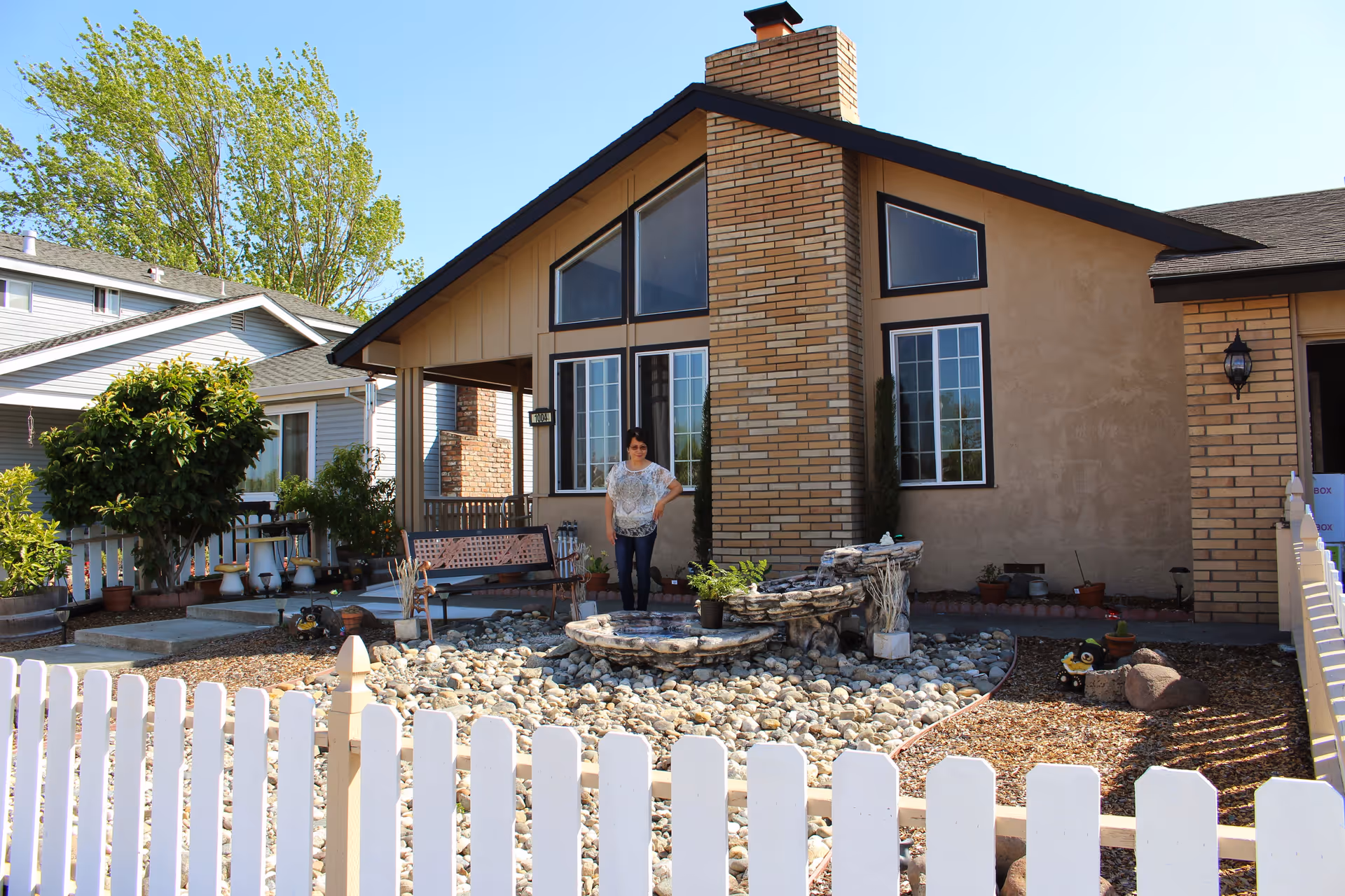 A single-story house with a brick chimney and large windows. There is a white picket fence in the foreground, a rock garden with a small water feature, and a woman standing near the house. Trees and neighboring houses are visible in the background under a clear blue sky.