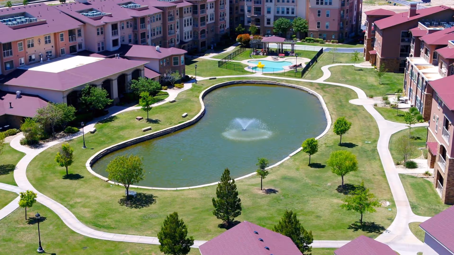 Aerial view of Isle at Raider Ranch senior living facility showing a landscaped courtyard with a large pond featuring a central water fountain, surrounded by walking paths, green lawns, trees, and multi-story residential buildings with red roofs.
