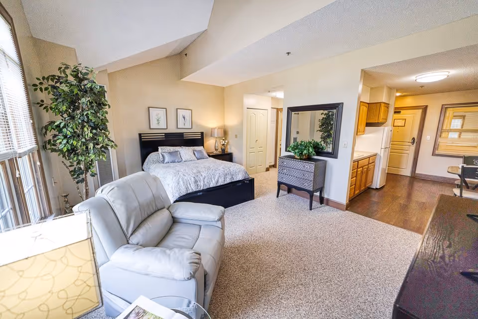 Studio-style senior living interior with a bed, recliner, dresser, and a small kitchenette visible.