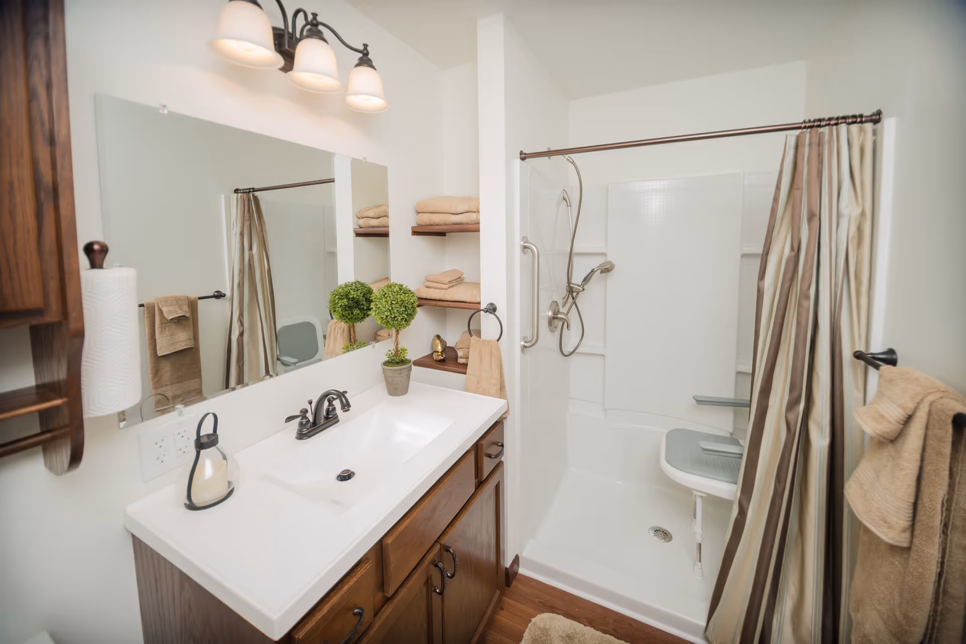 Well-lit bathroom with a wooden vanity and sink, large mirror, towel shelves, and a walk-in shower with a grab bar and shower seat.