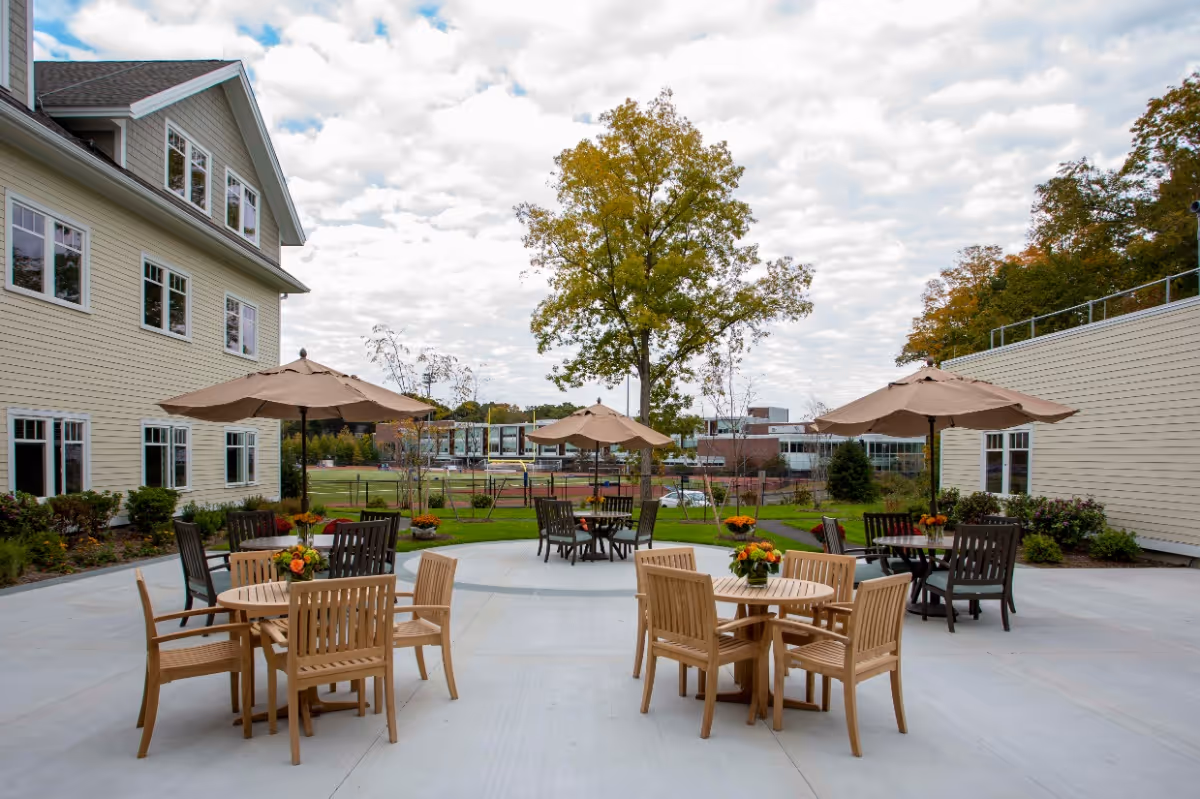 Outdoor patio area with several round wooden tables and chairs, each table adorned with a small flower arrangement. Large beige umbrellas provide shade over some tables. The patio is surrounded by buildings and greenery, with a large tree in the background under a partly cloudy sky.