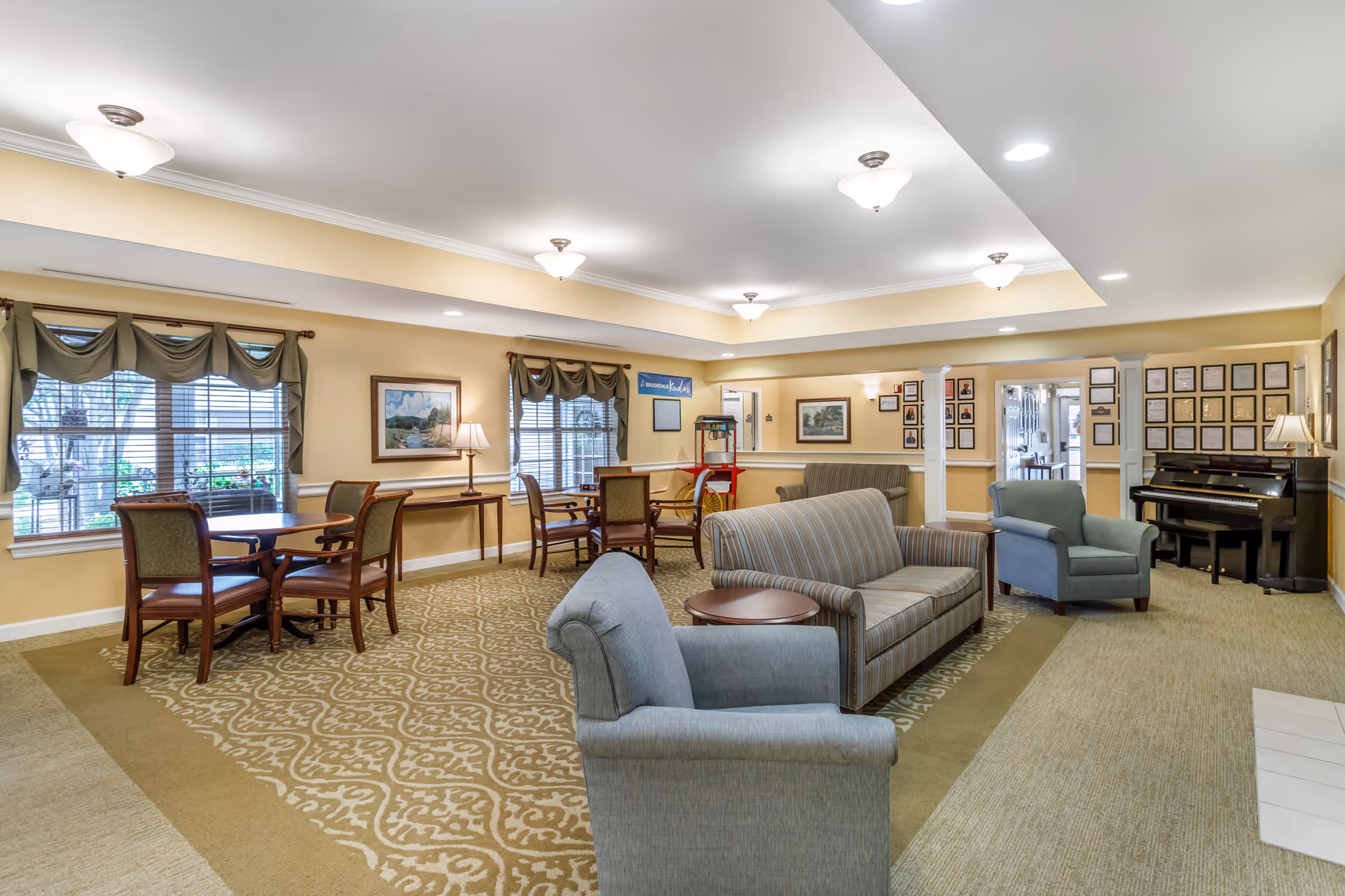 A spacious and well-lit common area in a senior living facility with beige walls and carpeted floors. The room features multiple seating options including a striped sofa, two blue armchairs, and several wooden tables with chairs. Large windows with green valances allow natural light to enter. A piano is placed against the far wall, and framed pictures and certificates decorate the walls. Ceiling lights provide additional illumination.