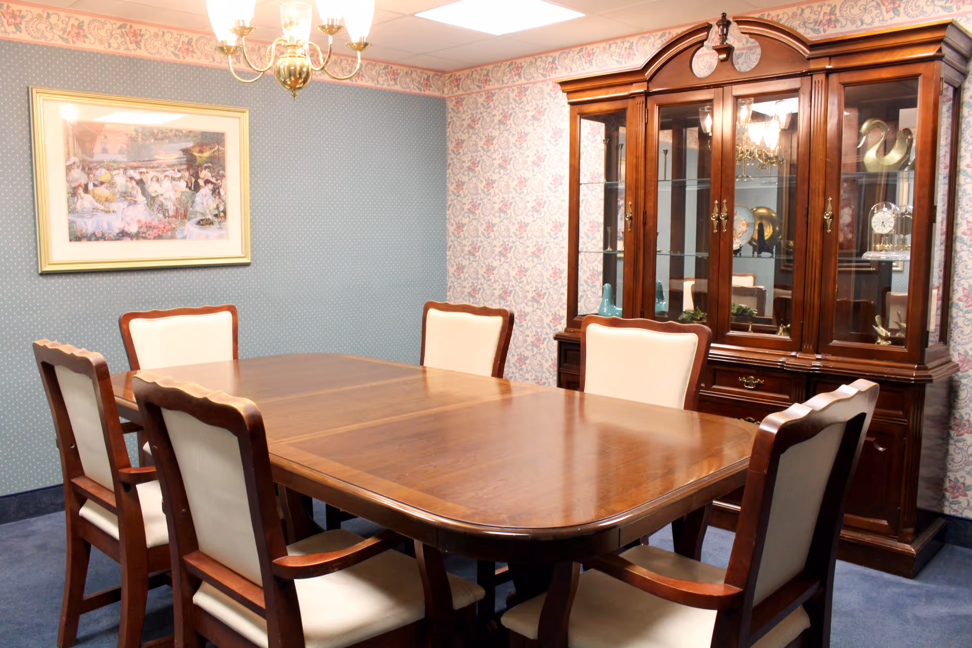 A traditional dining room with a large wooden dining table surrounded by six upholstered chairs. The room features floral and patterned wallpaper, a wooden china cabinet with glass doors displaying decorative items, a framed painting on the wall, and a brass chandelier hanging from the ceiling.