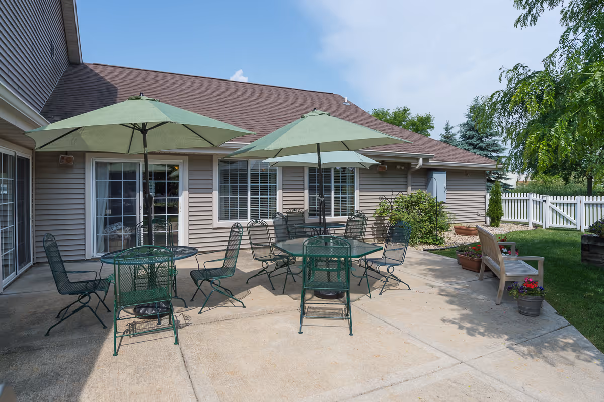 Outdoor patio area with two green metal tables, each with a green umbrella and several matching chairs. The patio is adjacent to a beige building with windows and sliding glass doors. There is a wooden bench with flower pots nearby, a white picket fence, and green grass and trees surrounding the area under a clear blue sky.