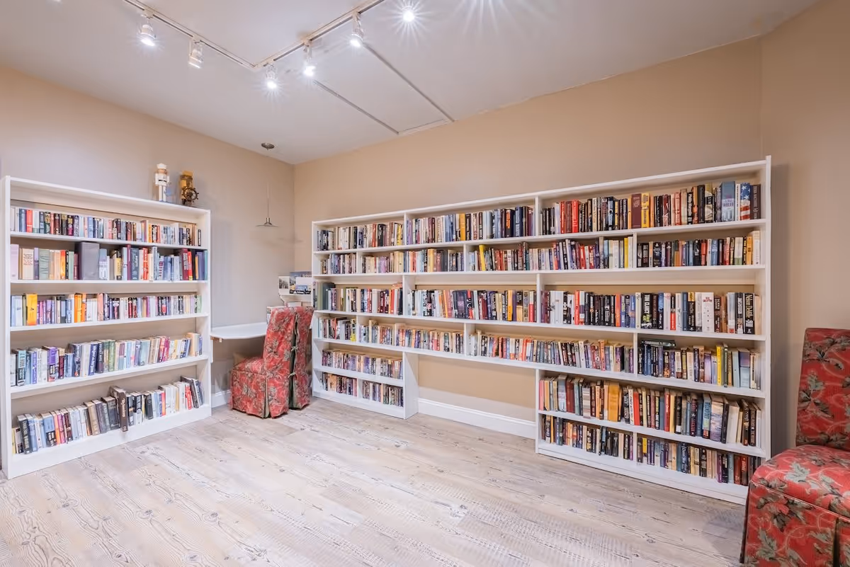 A cozy reading room with light wooden floors and beige walls, featuring white bookshelves filled with a variety of books. There are two red floral upholstered chairs and a small white corner desk with a hanging light above it. The ceiling has track lighting with multiple spotlights.
