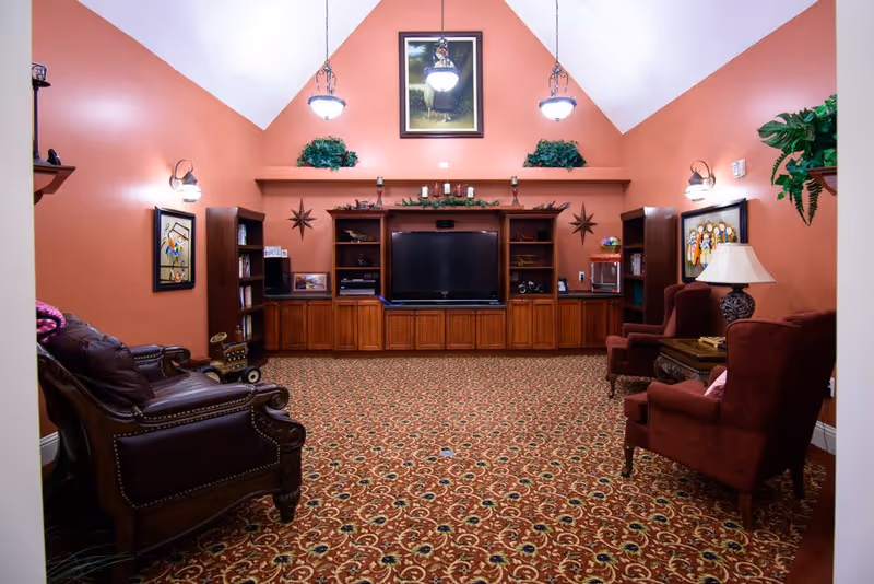 A cozy living room with patterned carpet, two red upholstered armchairs, a leather armchair, wooden cabinets with a large flat-screen TV in the center, wall-mounted lights, framed artwork, and decorative plants on a shelf above the TV.