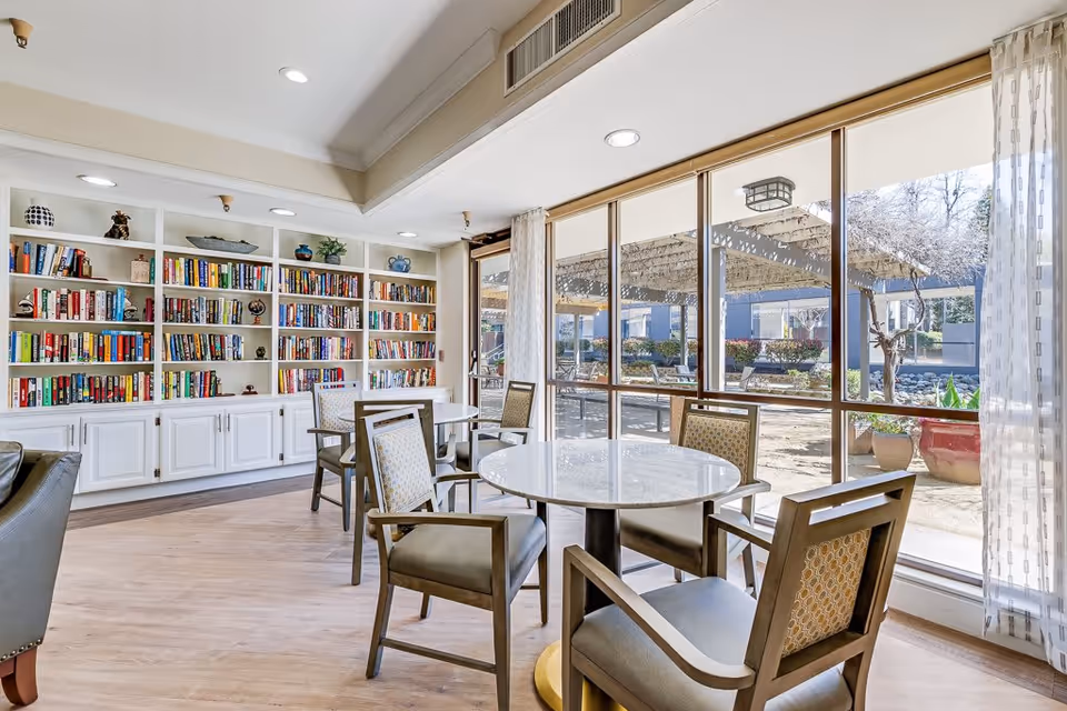 A bright and inviting common area in a senior living facility featuring round tables with chairs, a large built-in bookshelf filled with books and decorative items, and large windows that provide a view of an outdoor patio with pergolas and seating.