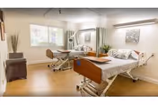 A bright and clean room with two hospital-style beds, each with white bedding and pillows. The room has light-colored wooden flooring, a large window letting in natural light, a small brown ottoman, and some wall decorations including a framed picture and a tall vase with dried plants.