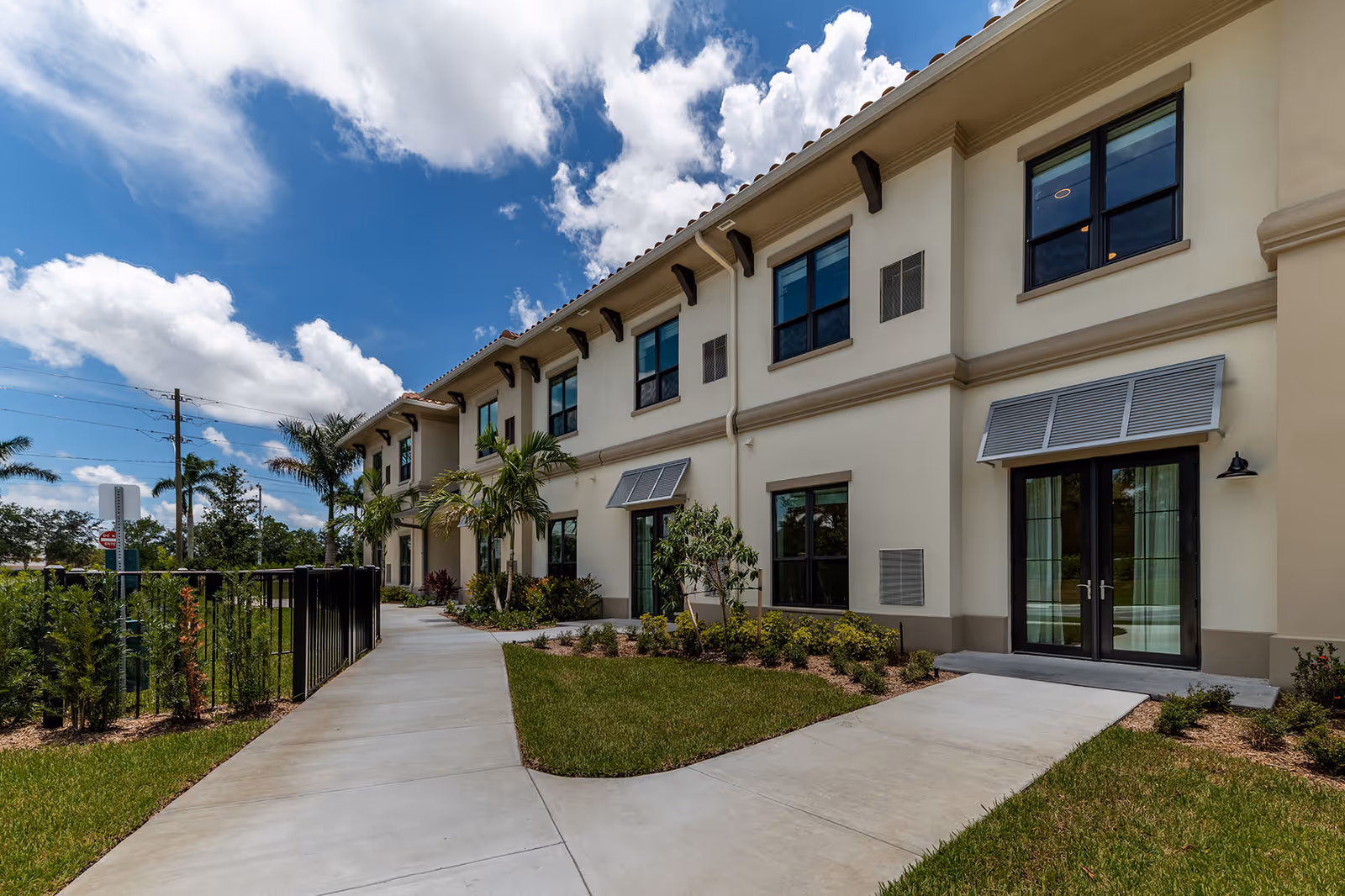 Exterior view of a two-story senior living facility building with beige walls, multiple windows, and glass doors. The building is surrounded by a well-maintained lawn, small shrubs, palm trees, and a concrete walkway leading to the entrance. The sky is partly cloudy with blue patches.