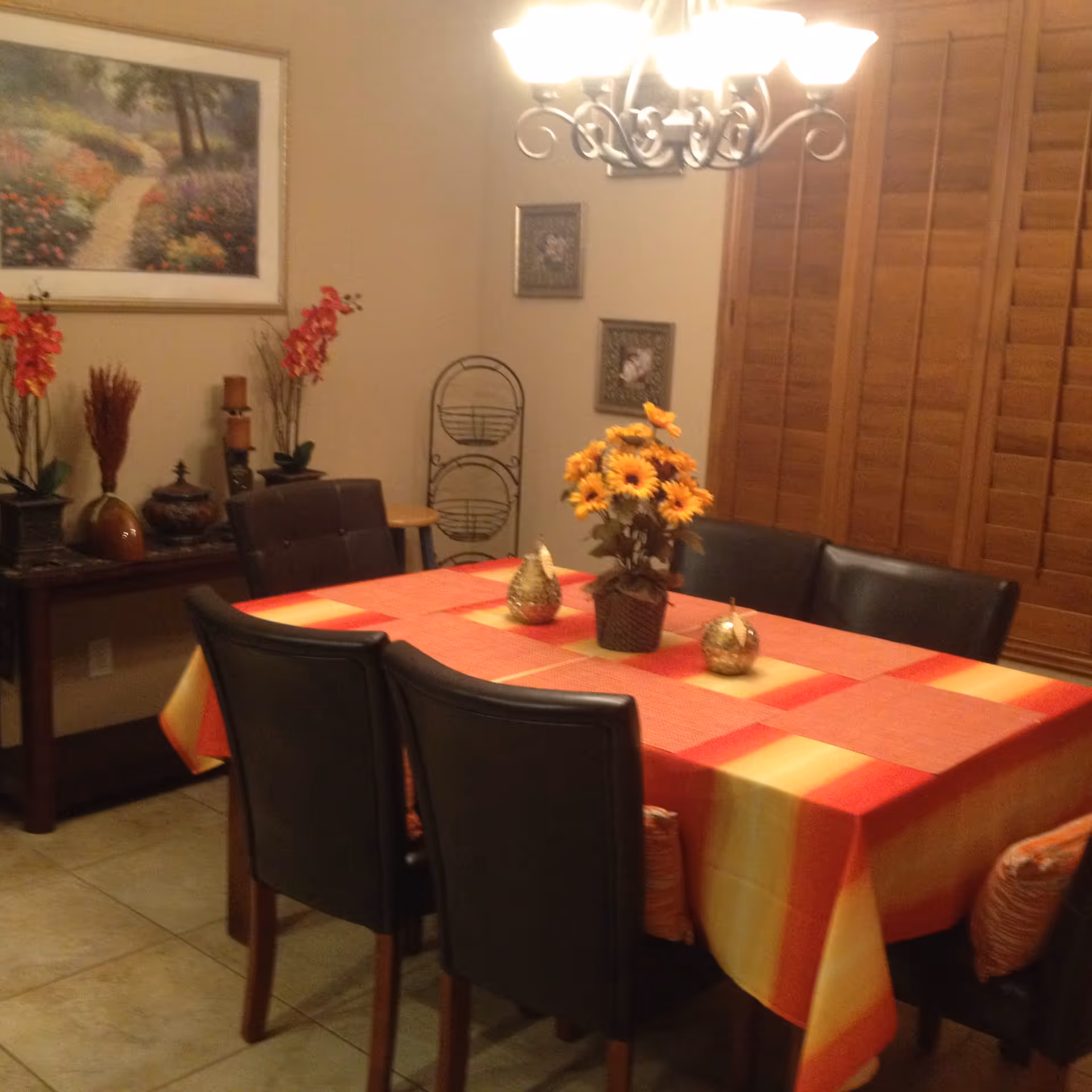 Dining room with an orange-and-red tablecloth on a rectangular table surrounded by dark chairs, a sunflower centerpiece, wall art, and wooden shutters.