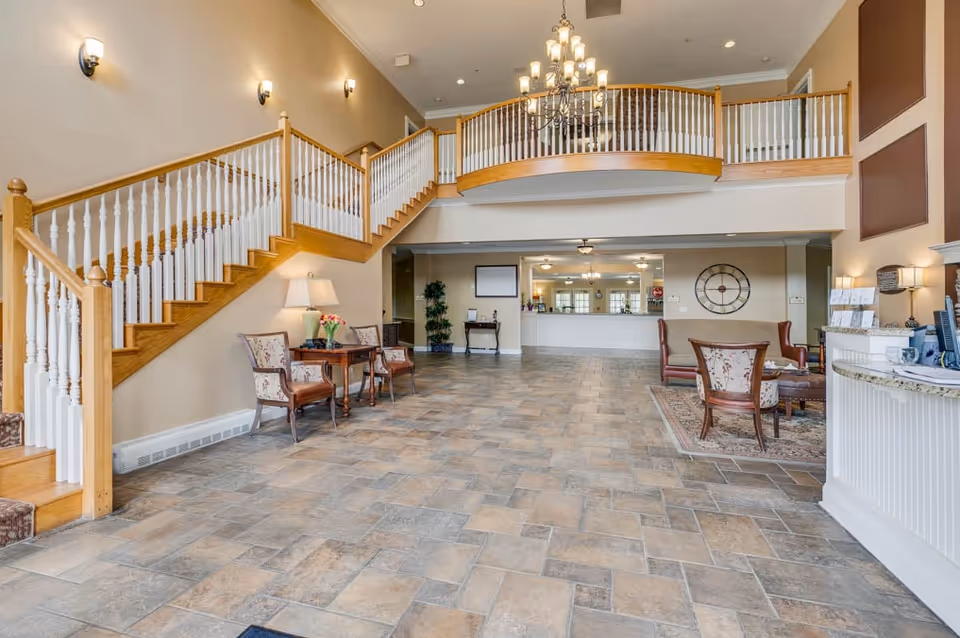 Spacious senior living lobby with tiled floor, seating areas, reception desk, and a wooden staircase with an upper balcony.
