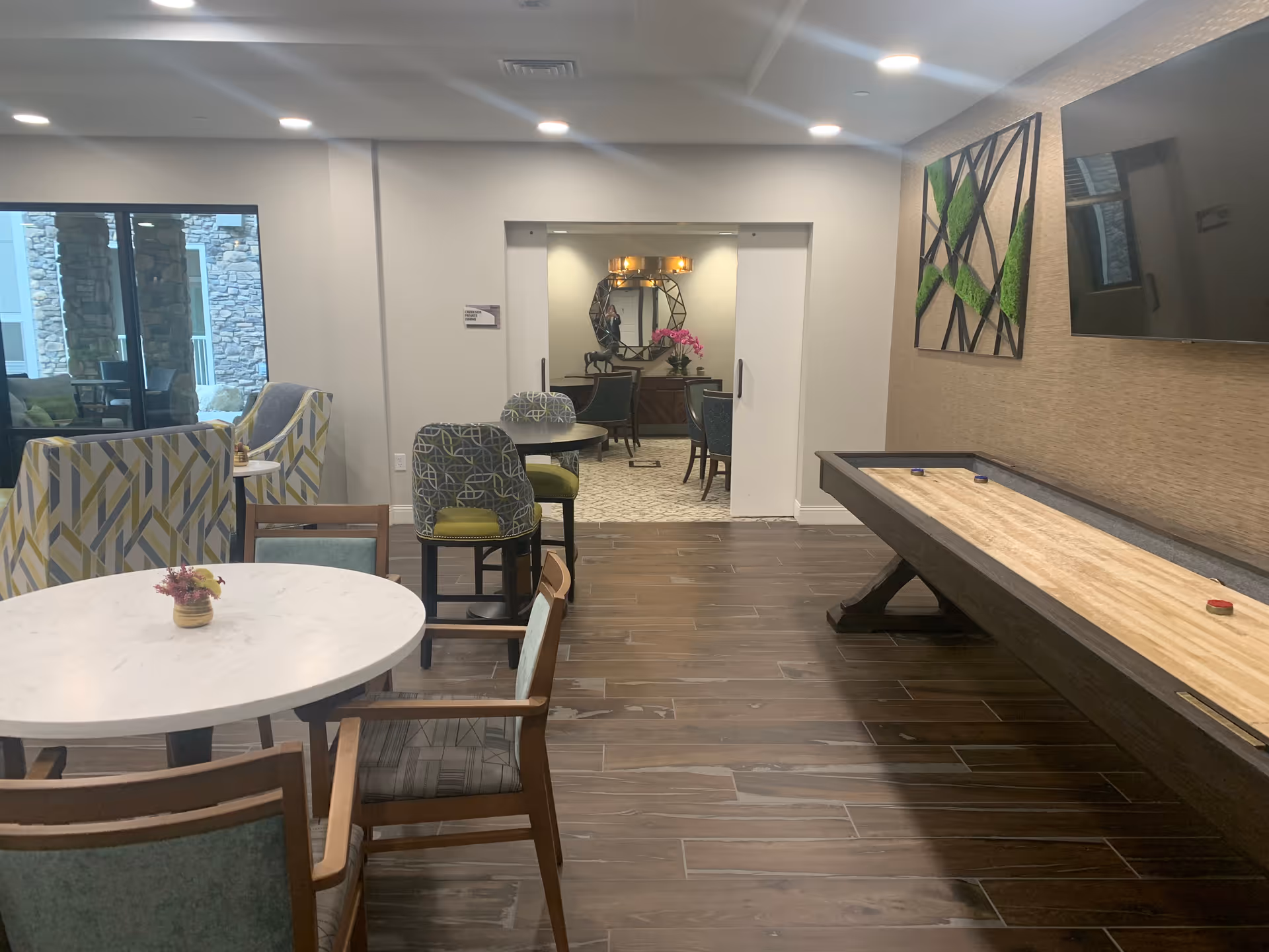 Community lounge with tables, chairs, patterned armchairs, and a shuffleboard table.