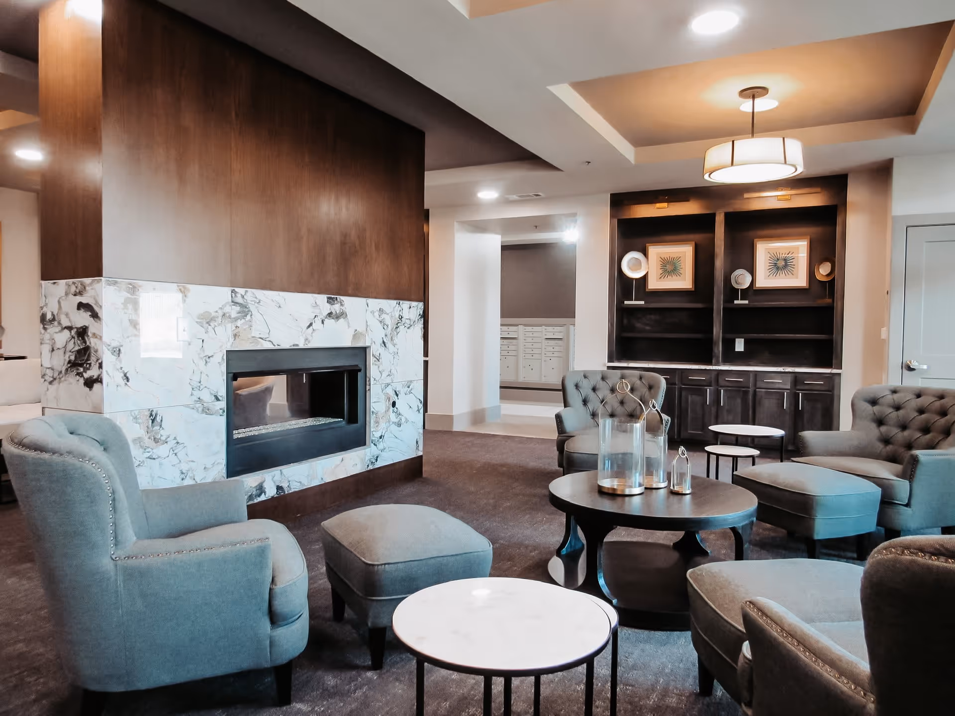 A cozy living room area in a senior living facility featuring a modern fireplace with marble surround, several upholstered armchairs and ottomans in a muted blue-gray color, round wooden coffee tables with decorative lanterns, and built-in dark wood shelving with framed artwork and decorative plates. The space has soft lighting and a neutral color palette.