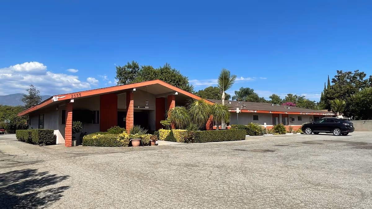 Single-story senior living facility with a covered red-trimmed entrance, landscaped shrubs, and a large parking lot under a clear blue sky.
