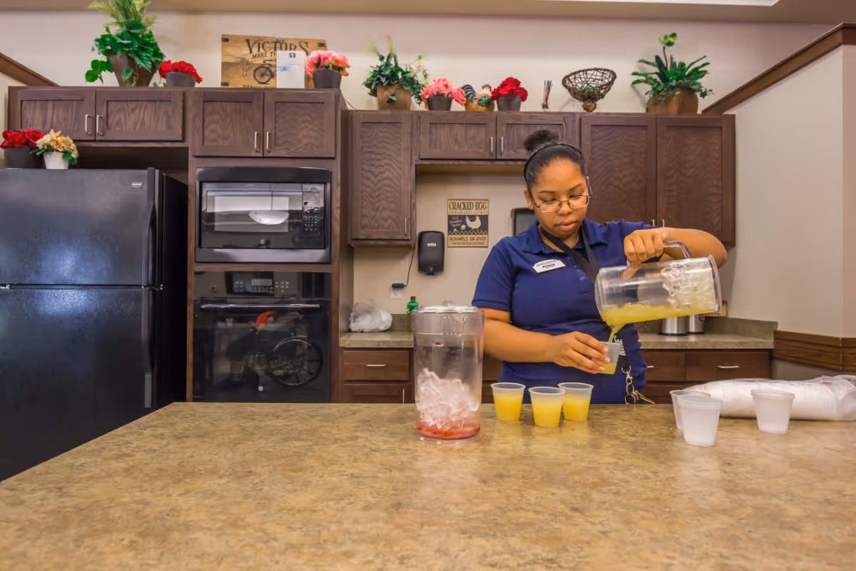 A woman wearing glasses and a navy blue shirt is pouring yellow juice from a large pitcher into small plastic cups on a kitchen counter. The kitchen has dark wooden cabinets, a black refrigerator, a microwave, and an oven. There are decorative plants and flowers on top of the cabinets.