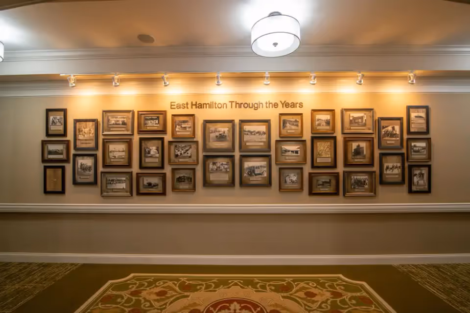 A wall display titled 'East Hamilton Through the Years' featuring multiple framed historical photographs arranged in a grid pattern, illuminated by track lighting above. The wall is painted beige with white trim, and a patterned carpet is visible on the floor in front of the display.