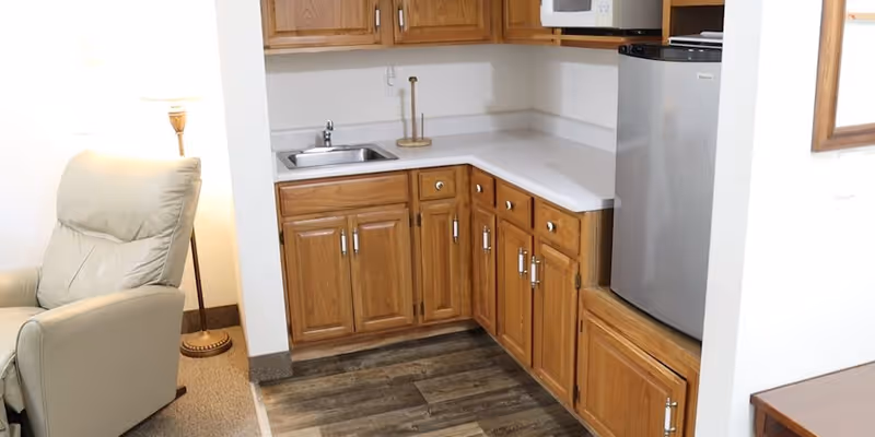 Small kitchenette with oak cabinets, a sink, countertop and mini-fridge beside a beige recliner and floor lamp.