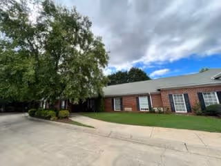 Exterior view of a single-story brick building with multiple windows, a green lawn, and a large tree near the driveway under a partly cloudy sky.