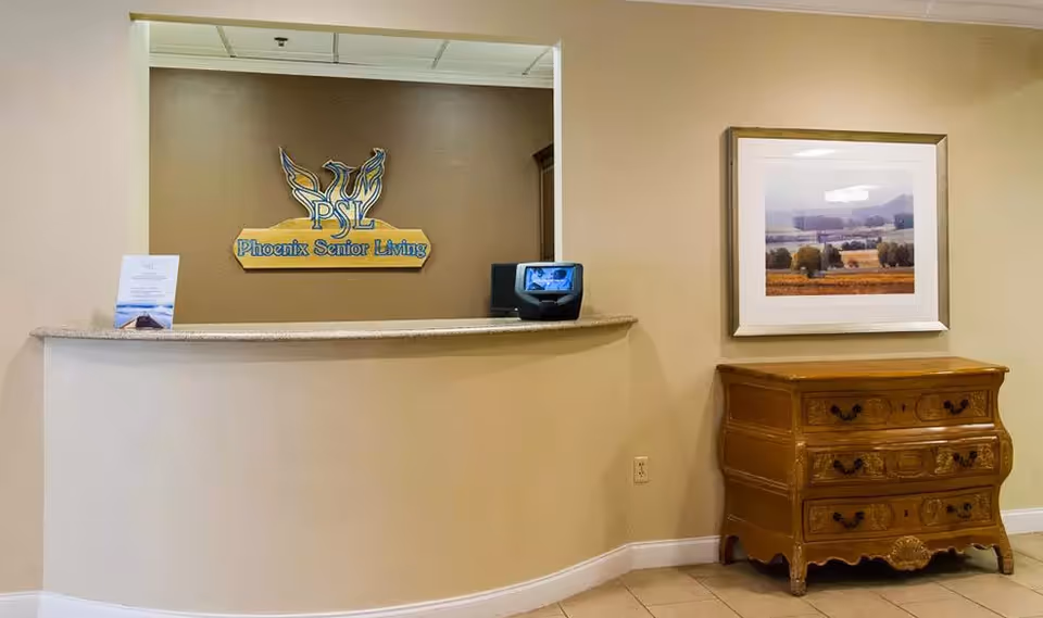 Reception area with a curved beige counter, a sign on the wall behind reading 'Phoenix Senior Living' with a phoenix logo, a small monitor on the counter, a framed landscape painting on the right wall, and a wooden chest of drawers beneath the painting.