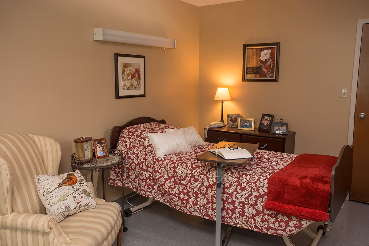 A cozy bedroom in a healthcare facility featuring a single bed with a red and white patterned bedspread and a red blanket at the foot. A small overbed table holds an open book and a pair of glasses. Next to the bed is a wooden dresser with a lamp and several framed photos. A beige striped armchair with a decorative pillow is positioned beside a small round side table with a clock and another framed photo. The walls are painted beige and decorated with two framed floral artworks.