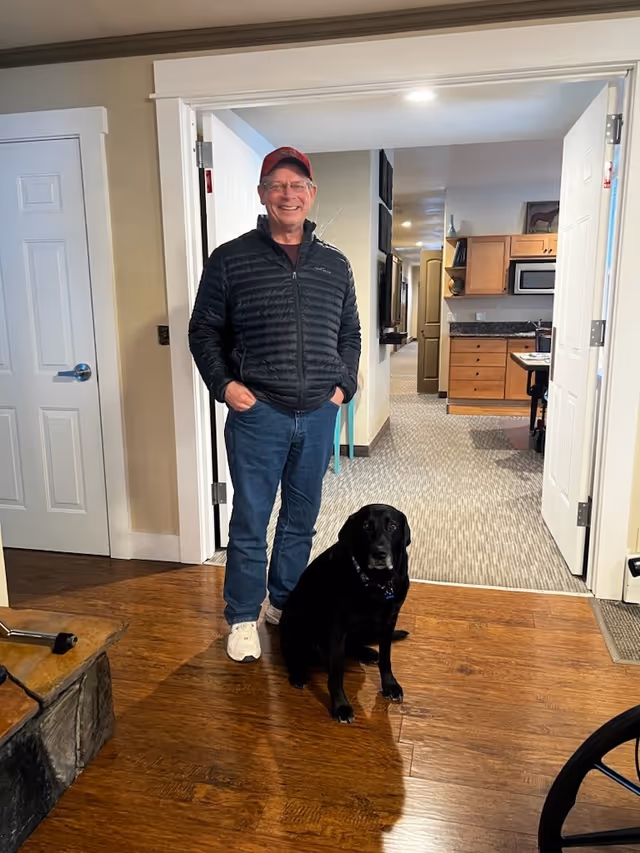 An elderly man wearing a red cap, glasses, a black jacket, blue jeans, and white sneakers stands smiling with his hands in his pockets inside a home. A black dog sits on the wooden floor next to him. Behind them is a hallway leading to a kitchen area with wooden cabinets and a microwave.